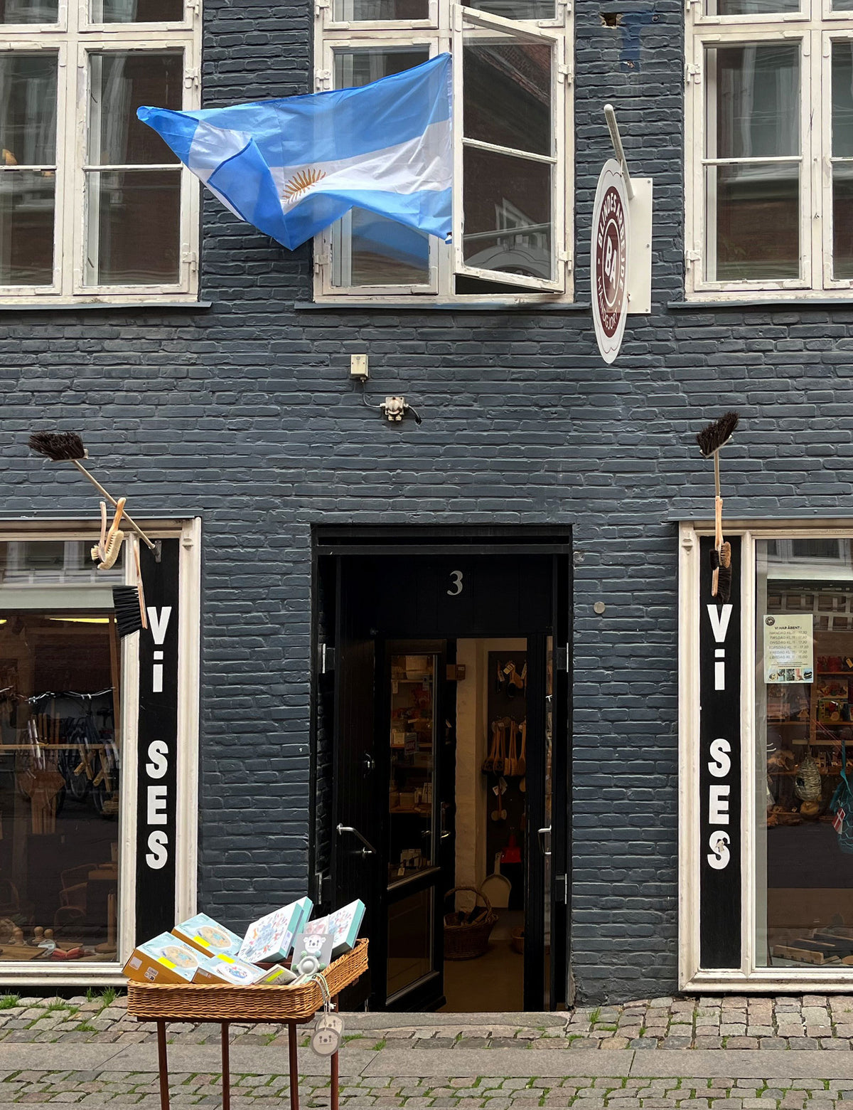 Mørk facade med “Vi Ses” i vinduerne, dannebrogsflag og en kurv med varer ved døren