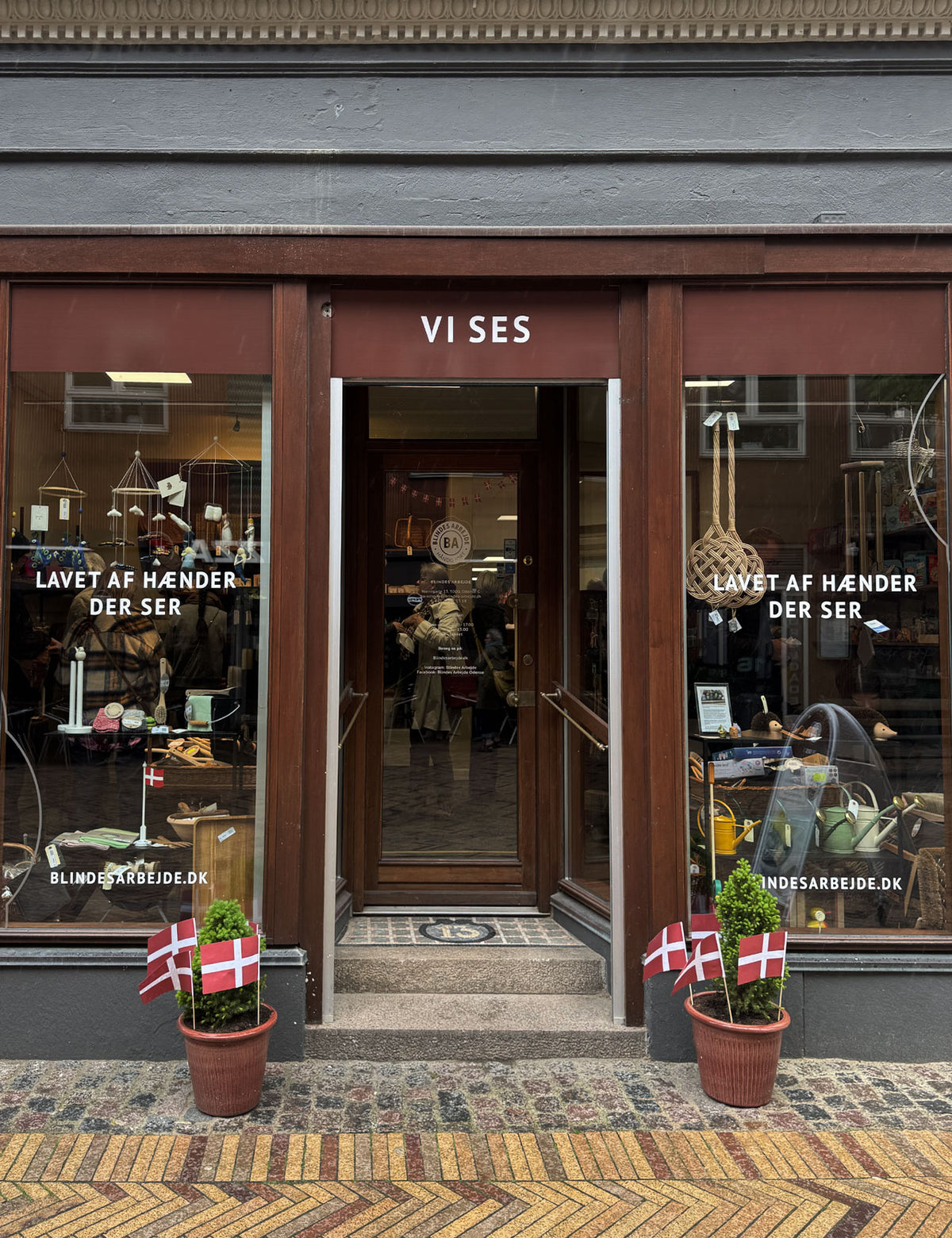 Butiksfacade i træ med “Vi Ses” over døren, dannebrogsflag og potteplanter på trappen