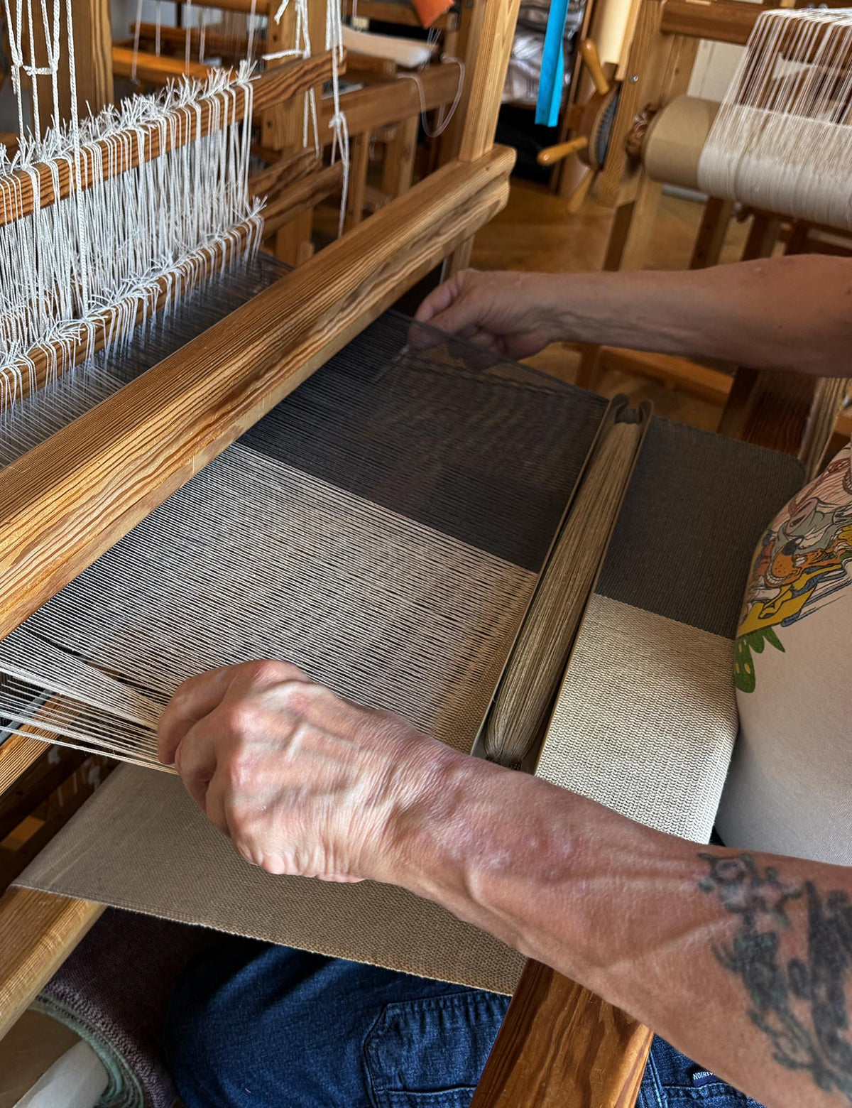 Hænder væver på en stor trævæv. Stoffet har brede striber i gråt og lys beige, og en træskytte med garn ligger klar i skurret.