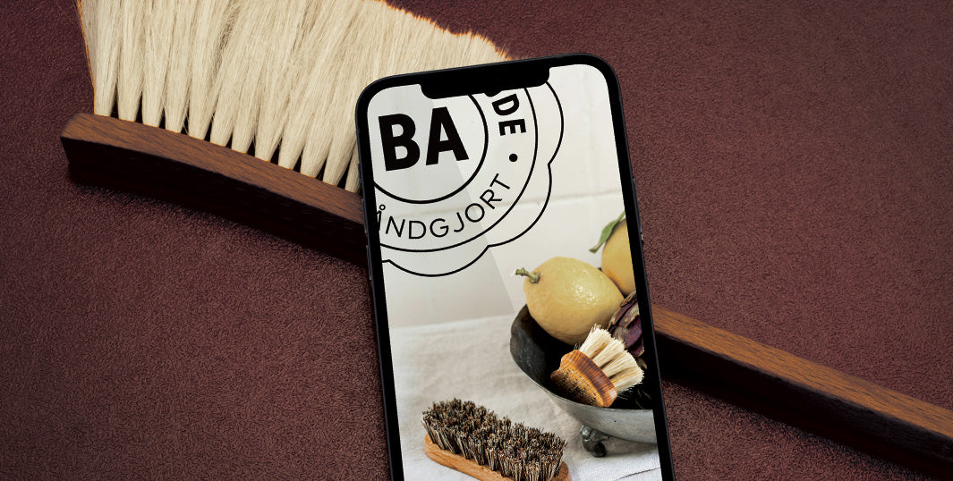 Mobiltelefon viser Blindes Arbejdes hjemmeside med BA-logo, citroner og børster. Ligger på rød bund ved siden af en kost.